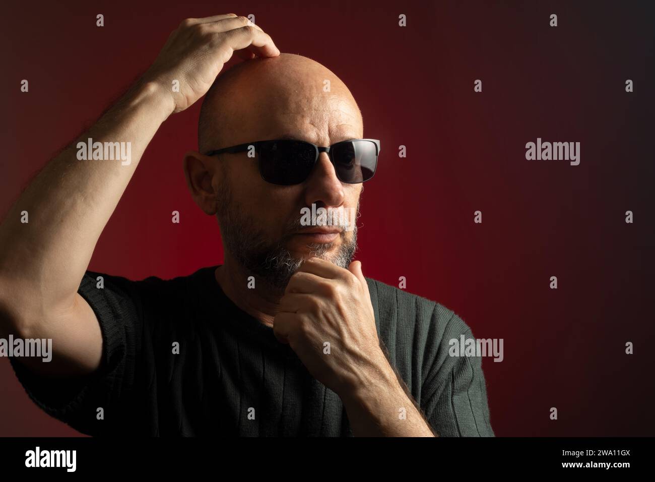 Caucasian man, bald, wearing sunglasses, confidently serious with his hand on his head and chin. Isolated on red background. Stock Photo