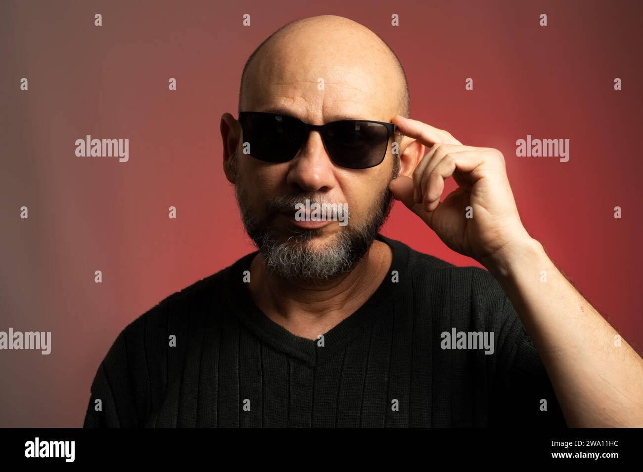 White, bald man, wearing sunglasses, confident and serious looking towards the camera. Isolated on red background. Stock Photo