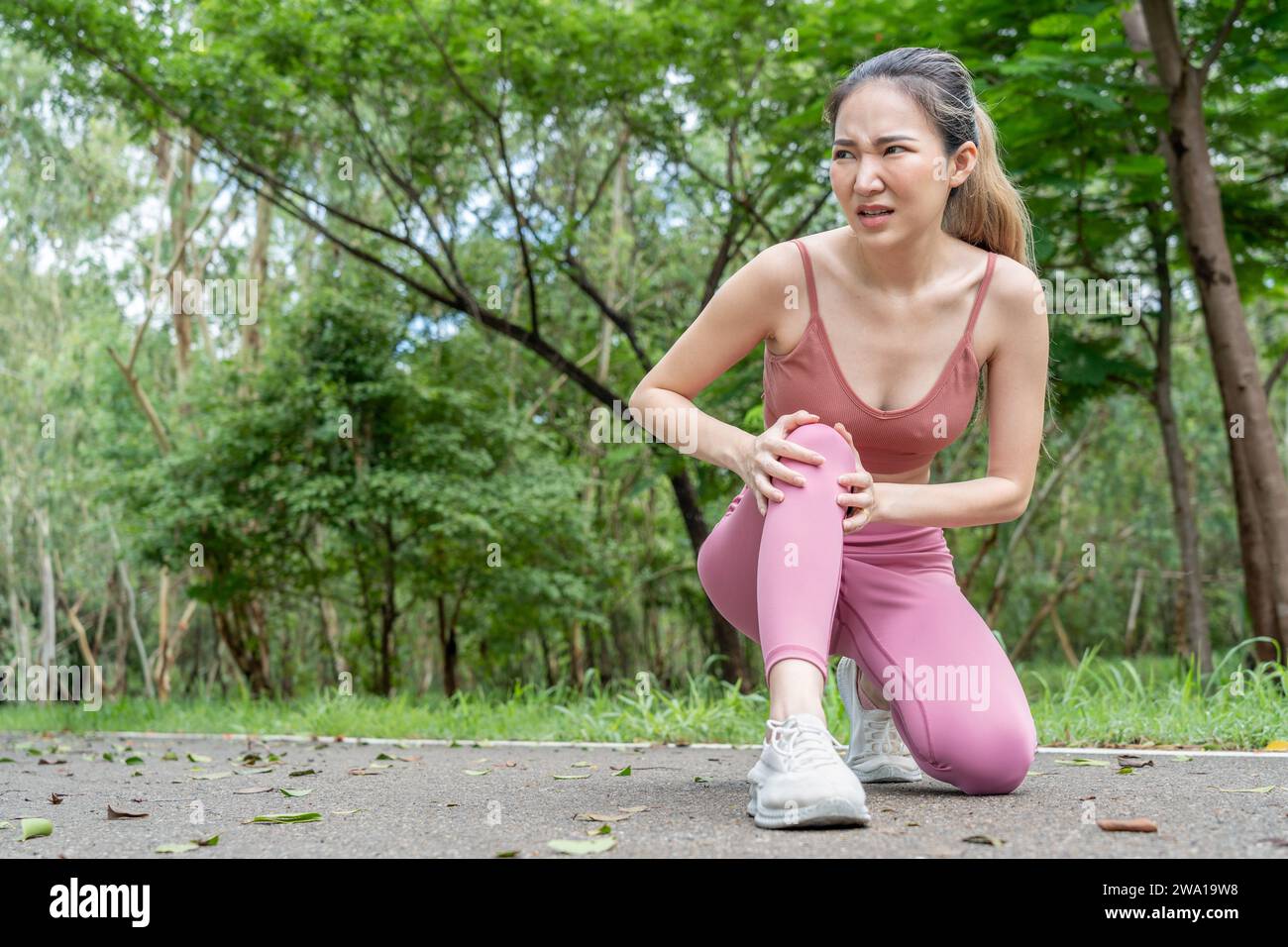 Young atractive Asian woman in fitness clothes with knee pain putting her hands over her troubled right knee while sitting down at a running track of Stock Photo