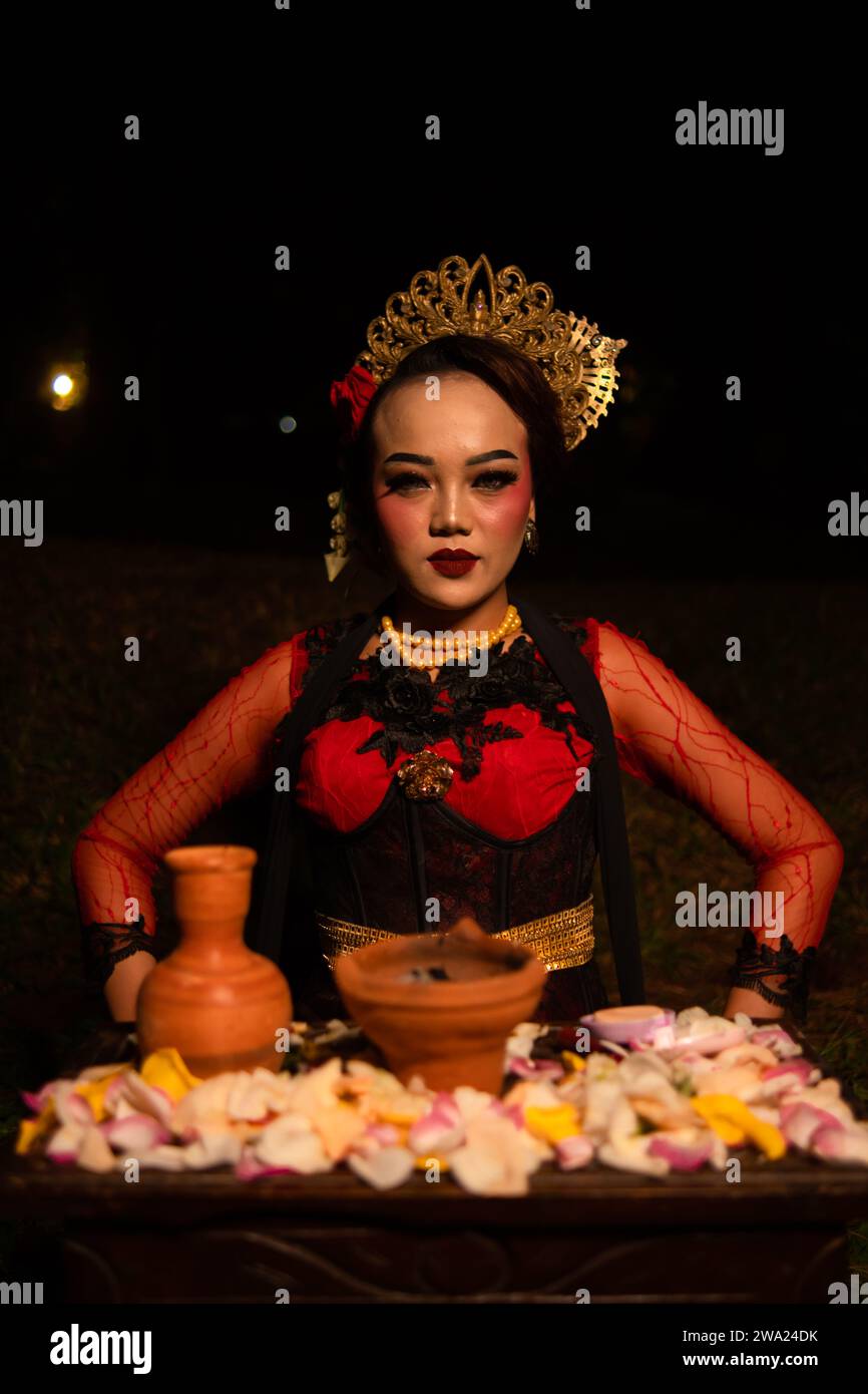 An Asian woman in a red dancer's dress is sitting at a ritual offering table full of flowers in a hut at night Stock Photo