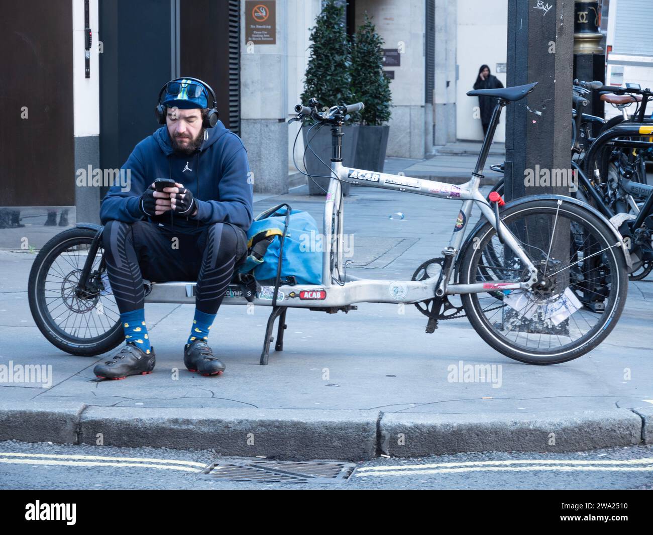 London, UK , cycle Courier,  Cargo bike messenger waiting for jobs in Soho, London, looking at mobile phone while seated on  bike in street Stock Photo