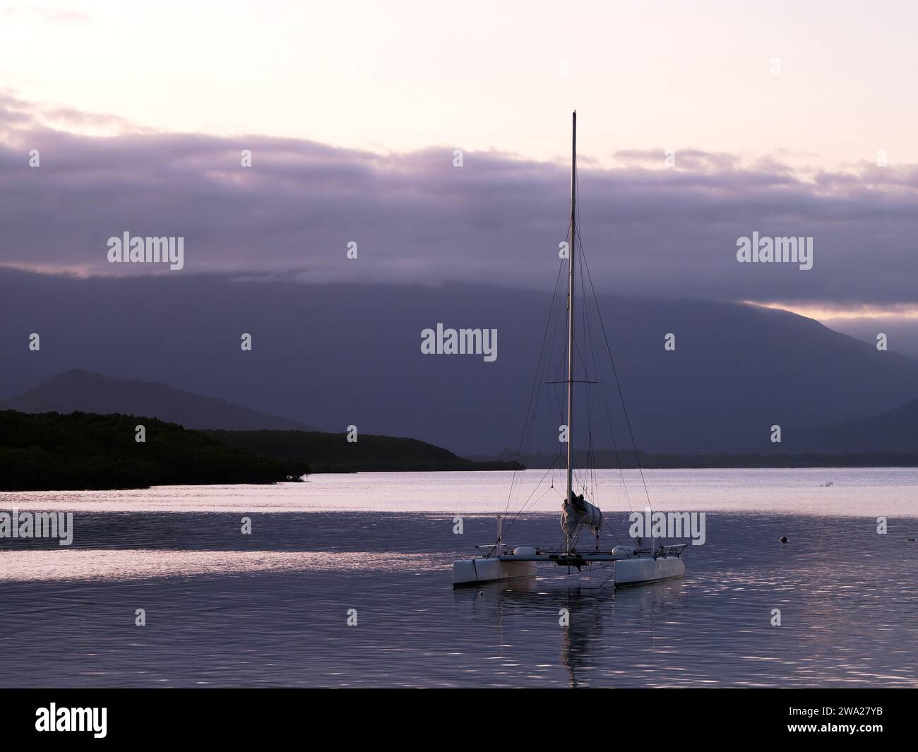 View of the sun setting over the distant hills and a catamaran moored in the foreground in Port Douglas Queensland Australia Stock Photo