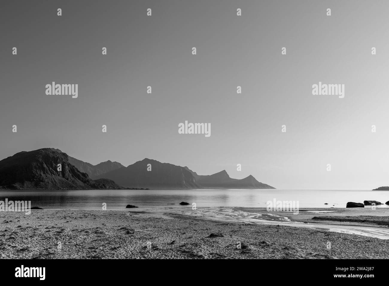 The grayscale palette of this image highlights the tranquil morning at Haukland Beach, with gentle waters meeting a layered mountain skyline. Lofoten Stock Photo