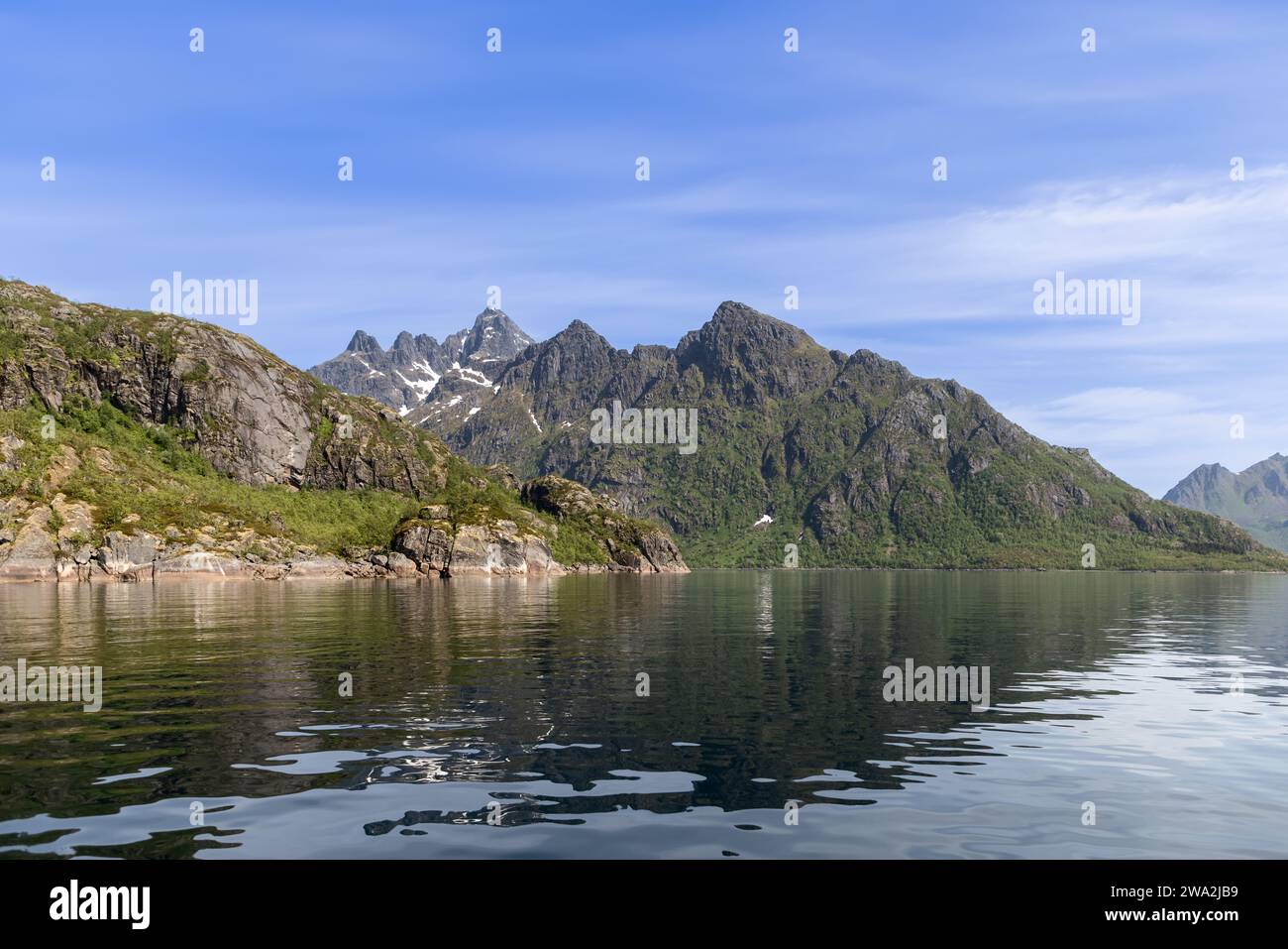 The mirror-like waters of Trollfjorden reflect the grandeur of the Lofoten peaks, with snow patches accenting the mountain's rugged green slopes Stock Photo