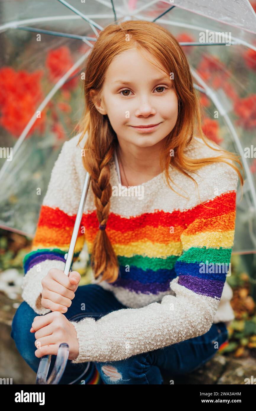 Outdoor spring portrait of adorable redheaded kid girl holding transparent umbrella, wearing rainbow pullover, child playing outside on a rainy day, f Stock Photo