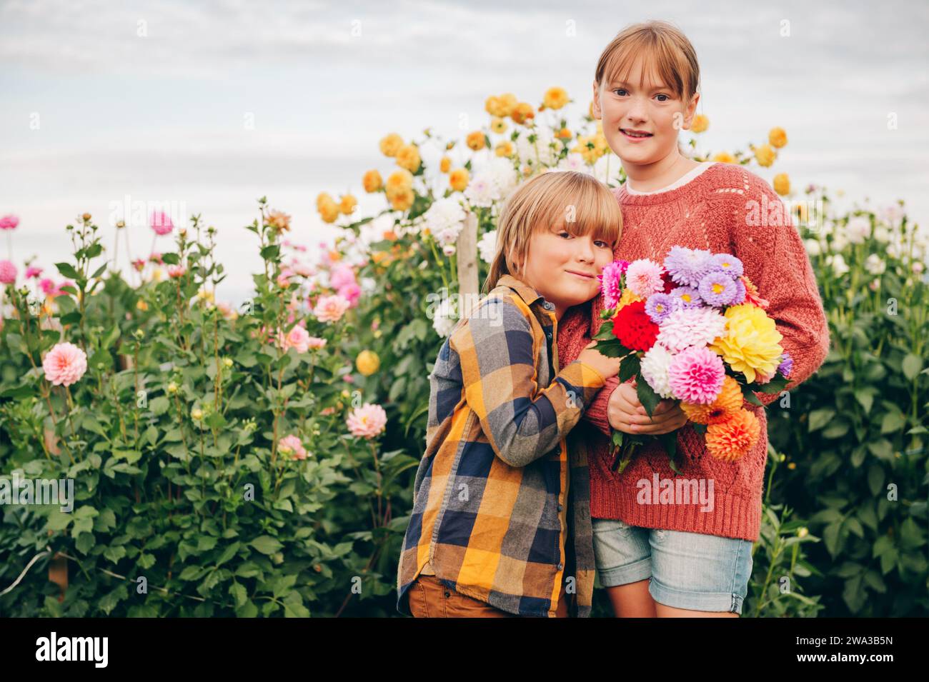 Outdoor portrait of two funny kids plying in autumn garden, holding dahlia flowers bouquet Stock Photo
