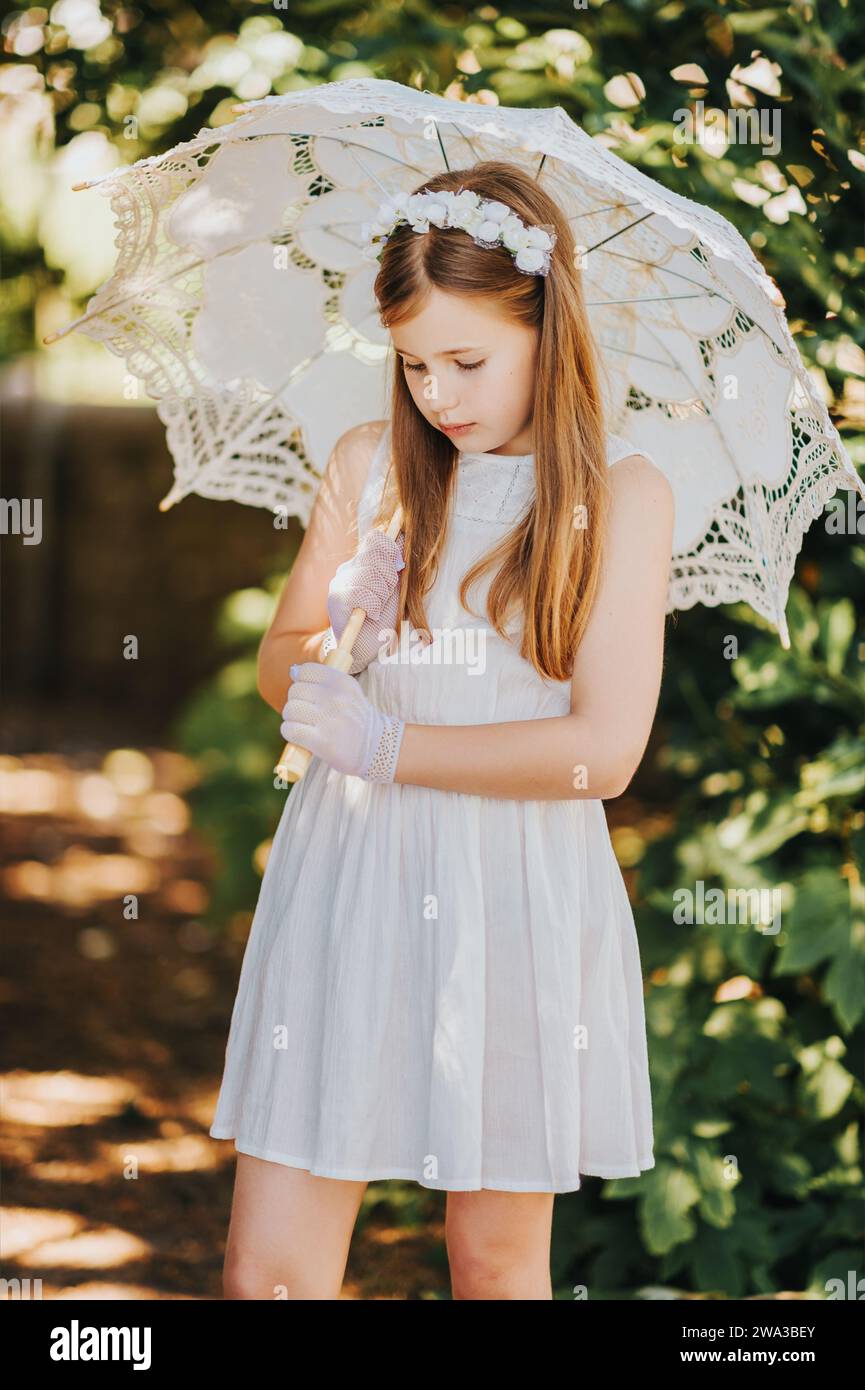 Outdoor portrait of romantic little girl, wearing white dress, gloves, flower headband, holding lace umbrella Stock Photo