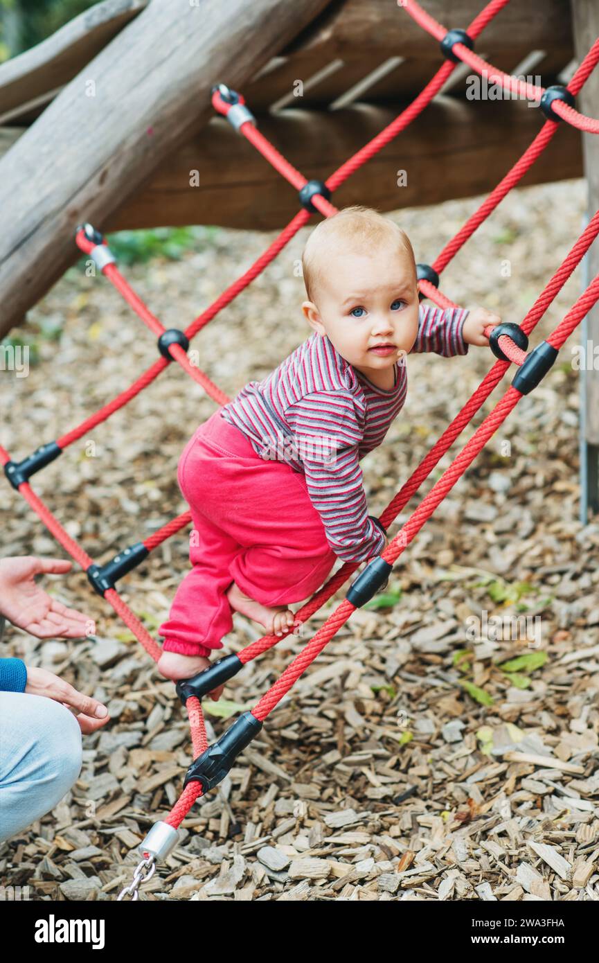 Adorable baby girl climbing the net on playground. 9-12 months old child having fun outdoors Stock Photo