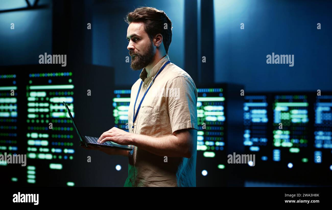 Programmer doing annual system examination, checking data on laptop of needed debugging in server mainframes housing advanced data storage hardware supporting important IT workloads Stock Photo