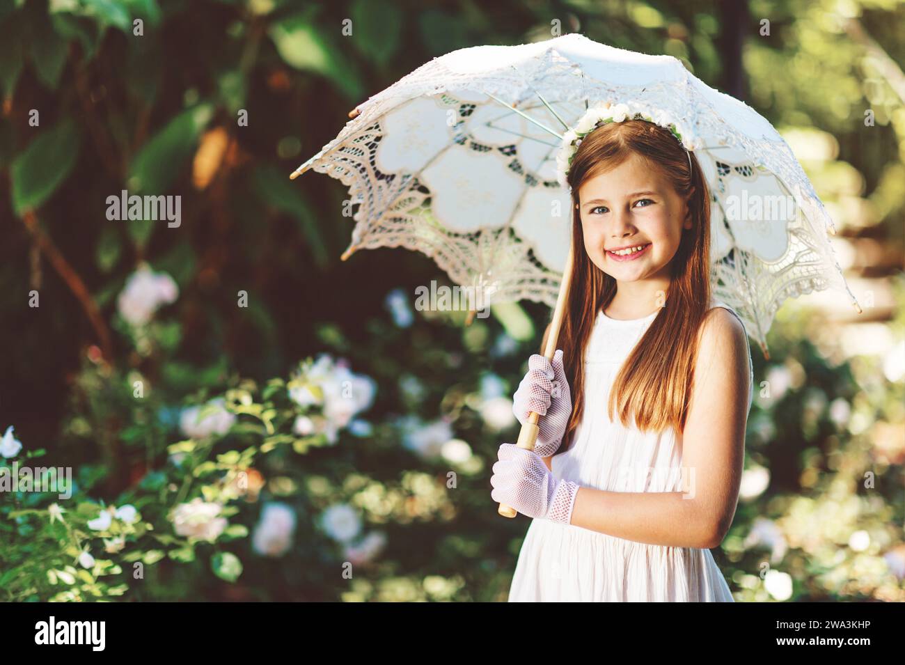 Outdoor portrait of romantic little girl, wearing white dress, gloves, flower headband, holding lace umbrella Stock Photo