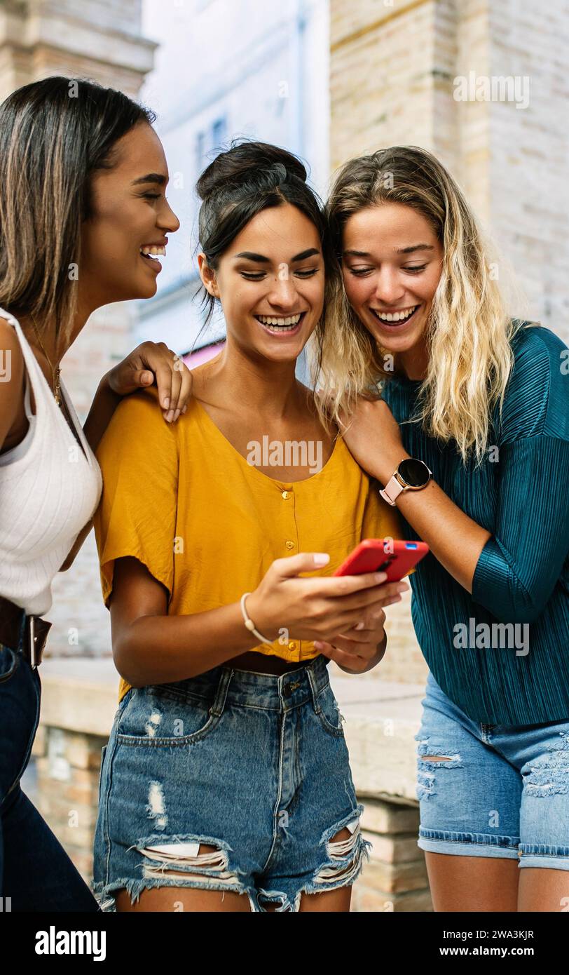 Multiracial young female friends laughing while using mobile phone outdoors. Stock Photo