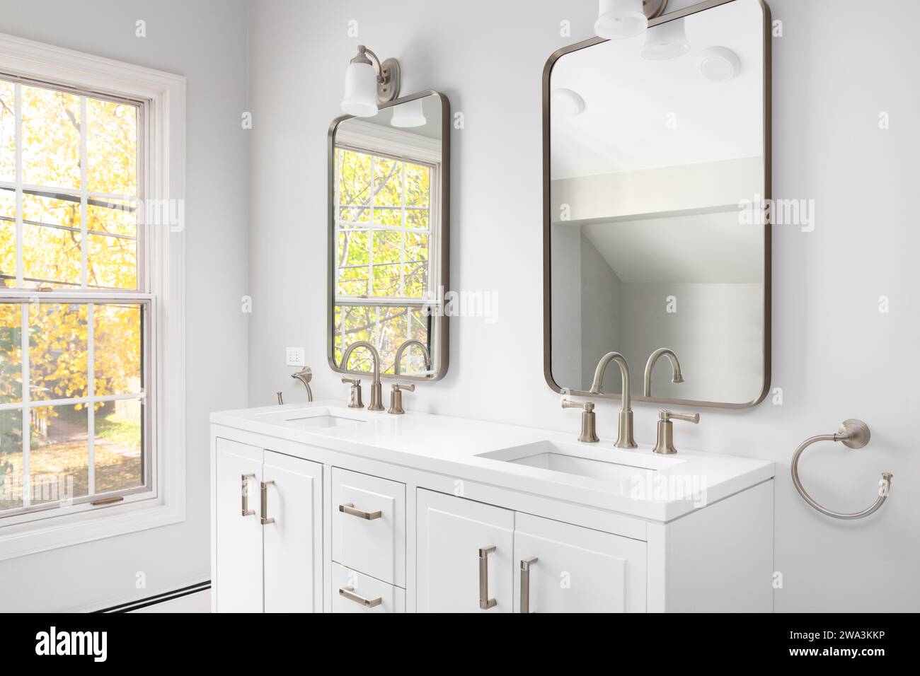 A bathroom detail with a white cabinet, bronze faucets and mirrors, and colorful trees out the windows. Stock Photo