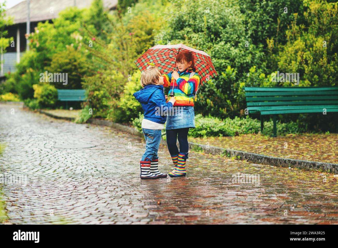 Two kids under umbrella, wearing rain boots and jackets Stock Photo