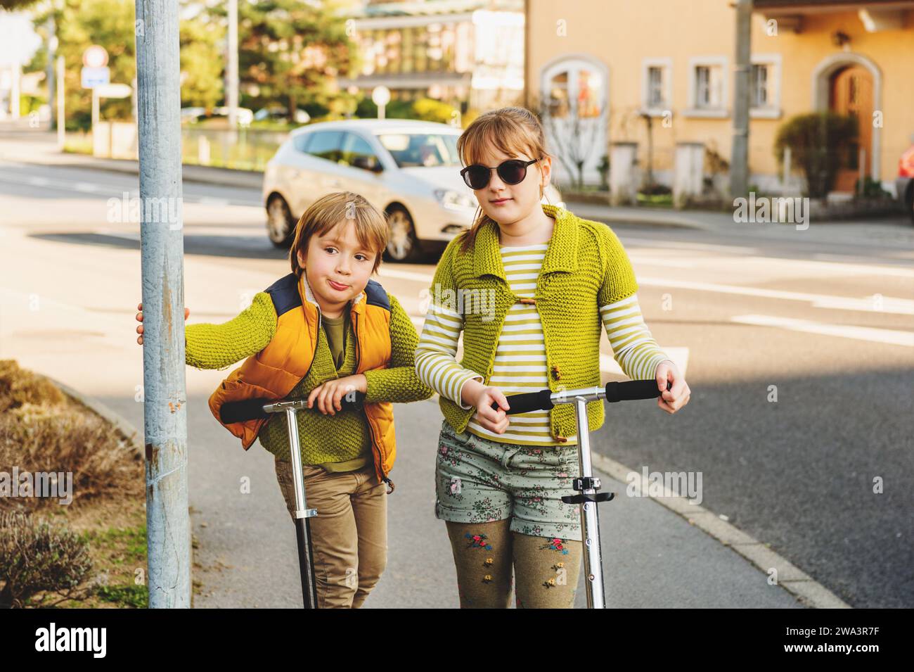 Two happy kids playing outdoors, fashion boy and girl posing on the street, riding scooters next to road Stock Photo