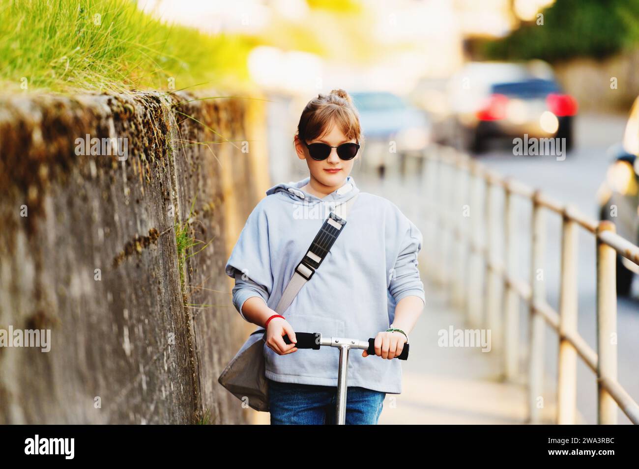 Happy 9 year old school girl with backpack in the streets of the city at sunset riding scooter, wearing sunglasses Stock Photo