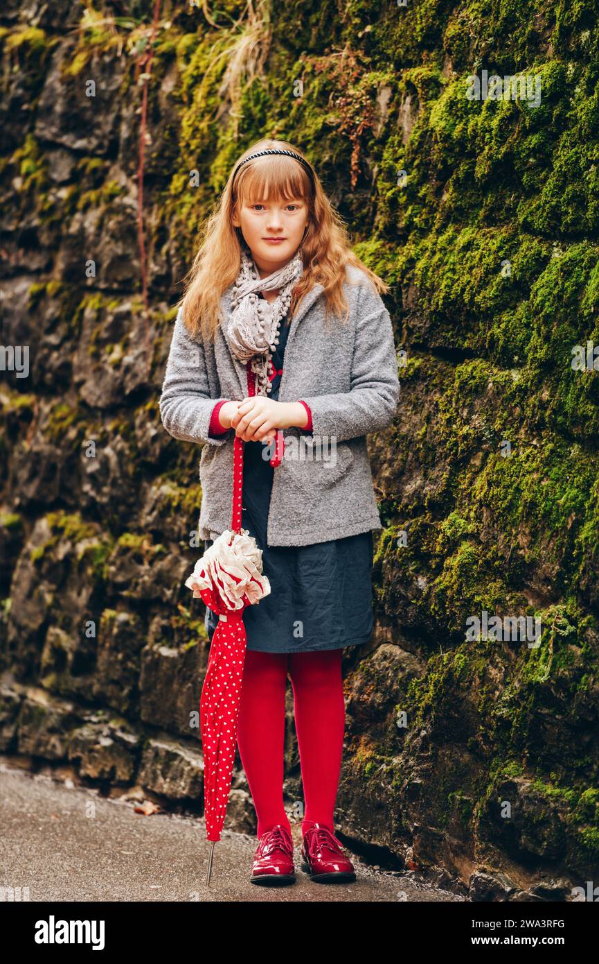 Outdoor portrait of pretty 9-10 year old little girl, wearing grey coat and scarf, holding red polka dot umbrella Stock Photo