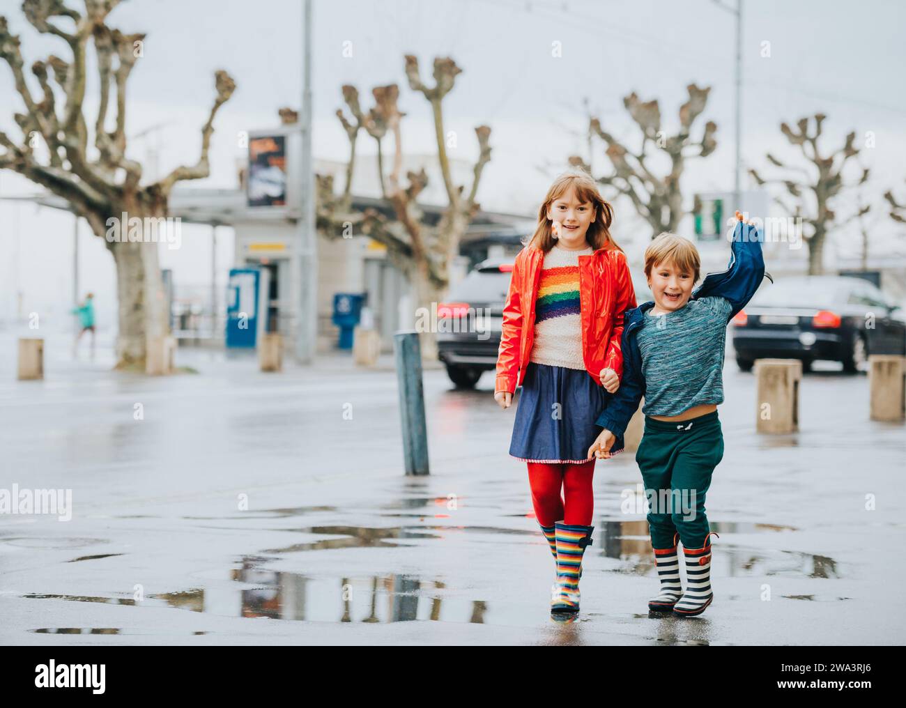 Two funny kids, little brother and sister playing together in a city under the rain in early spring. Chidren wearing rain jackets and boots Stock Photo