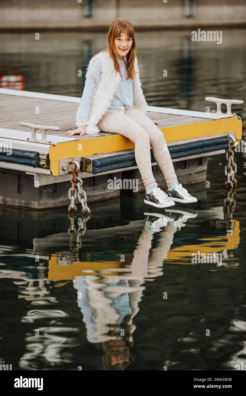 Outdoor fashion portrait of young preteen kid girl wearing white cap, faux fur gilet and silver mirror trainers, resting in a small port. Spring fashi Stock Photo