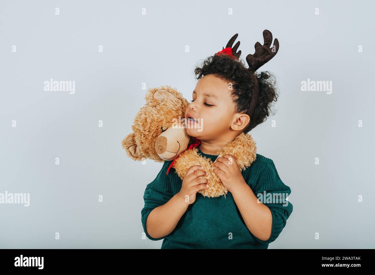 Christmas portrait of adorable toddler boy wearing green pullover, reindeer ears with Santa Claus' hat, holding teddy bear Stock Photo