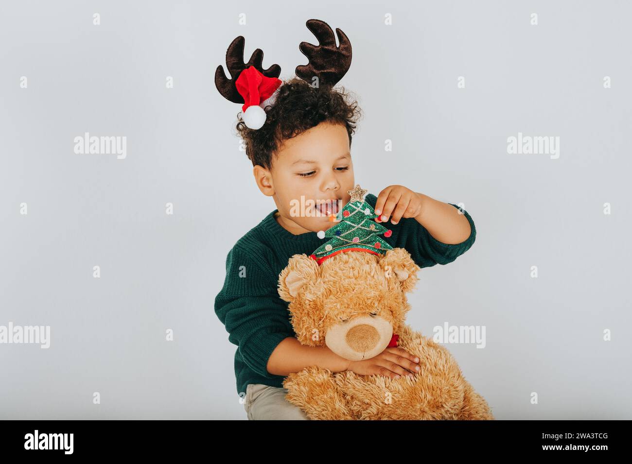 Christmas portrait of adorable toddler boy wearing green pullover, reindeer ears with Santa Claus' hat, holding teddy bear Stock Photo