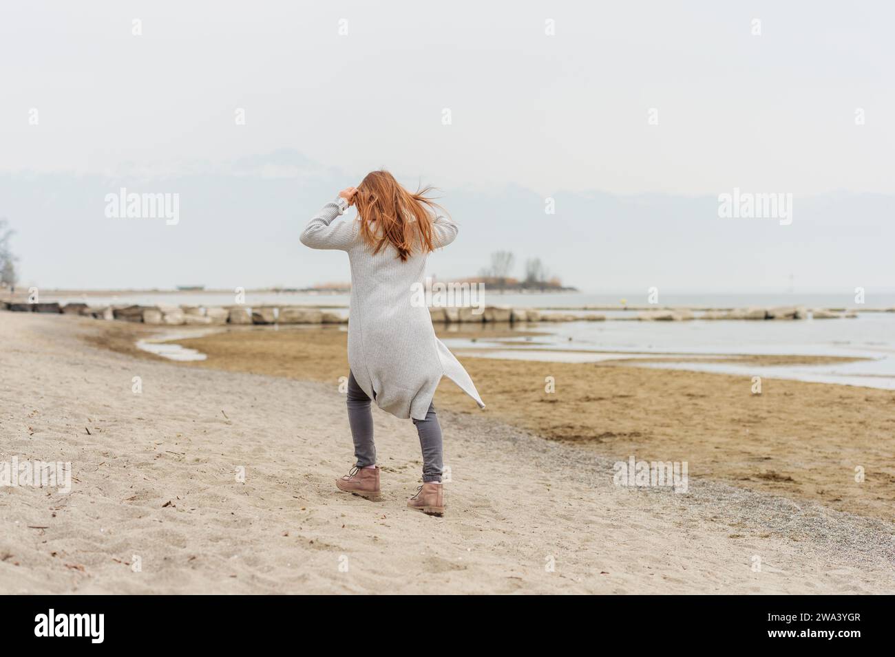 Adorable little girl of 8-9 years old playing by the lake, wearing grey trousers and long cardigan, back view Stock Photo