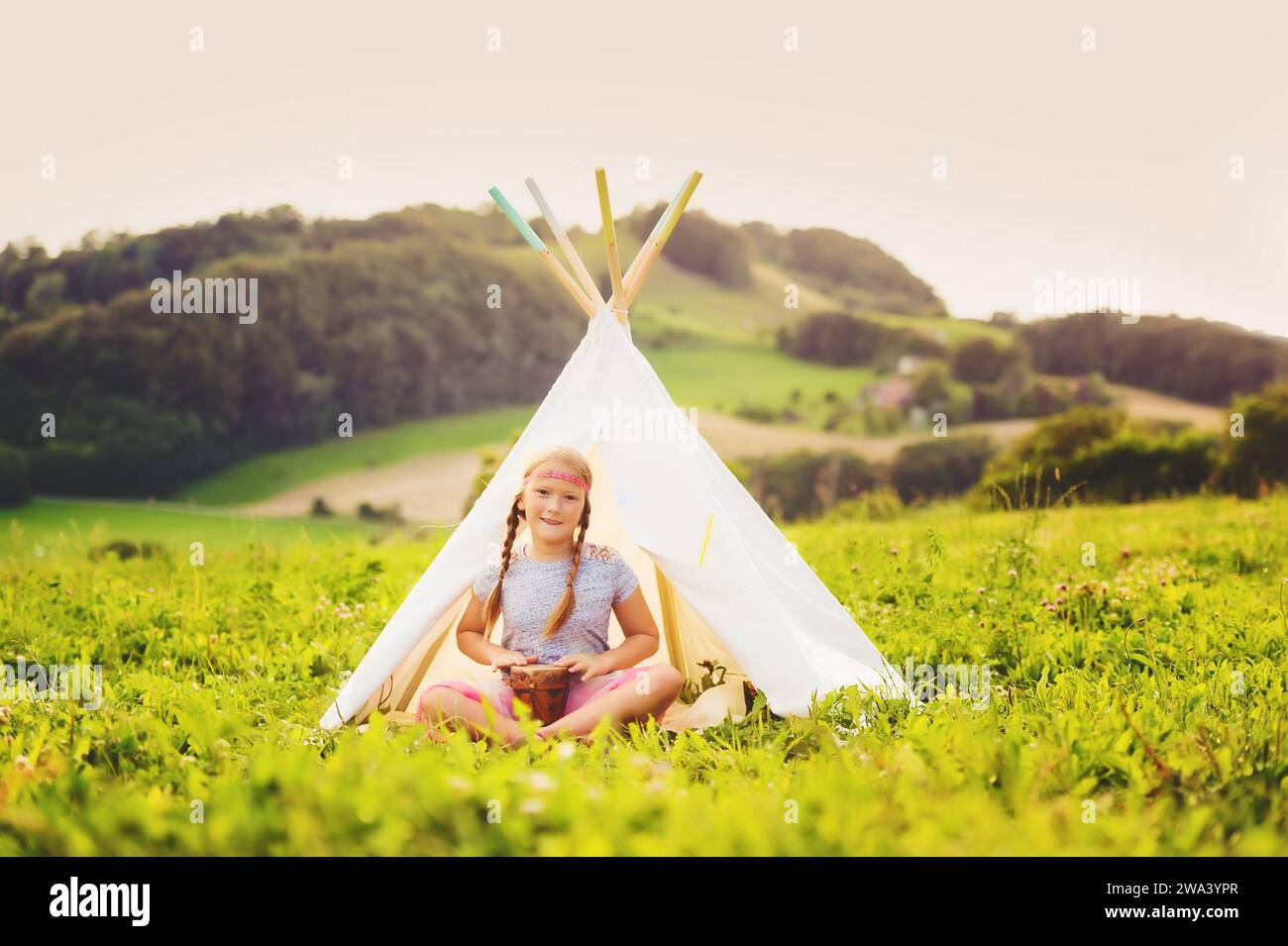 Cute little girl having fun outdoors, playing with tambour, sitting next to teepee Stock Photo