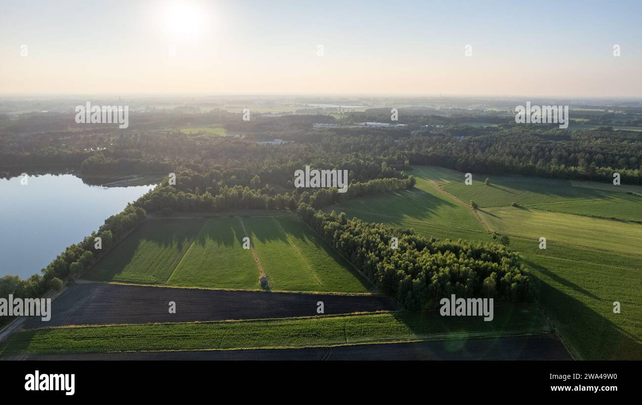 The photograph offers a sweeping view of the countryside at dawn, with the early sunlight bathing the fields, woods, and a tranquil lake in a soft, diffuse glow. The varying shades of green and the patterns of the agricultural landscape are clearly visible, presenting a tapestry of rural life awakening to a new day. Dawn's Early Light Over Countryside. High quality photo Stock Photo