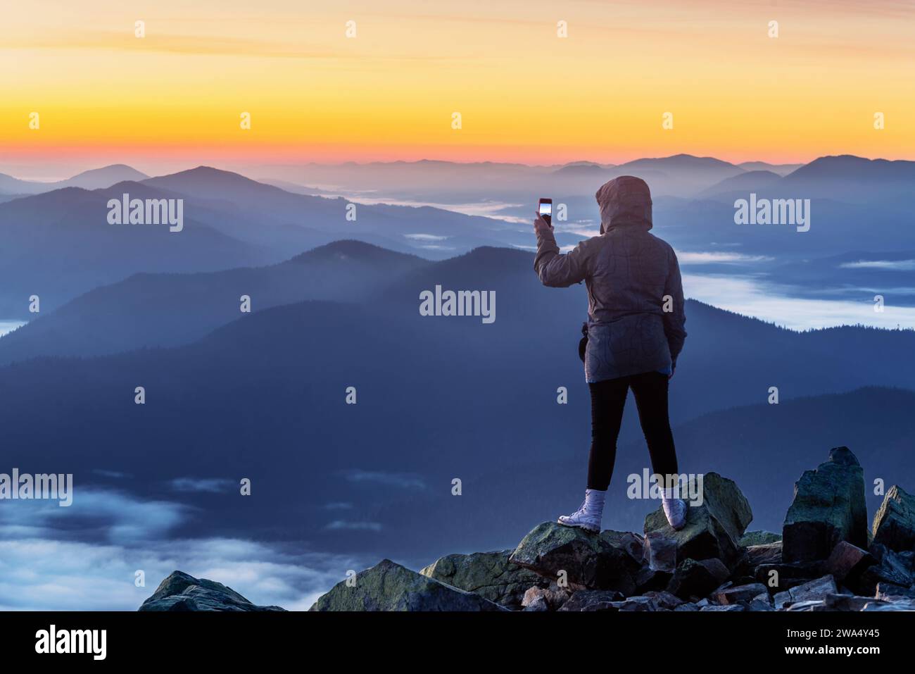 Young girl on the mountain peak taking photo with smart phone at sunset ...
