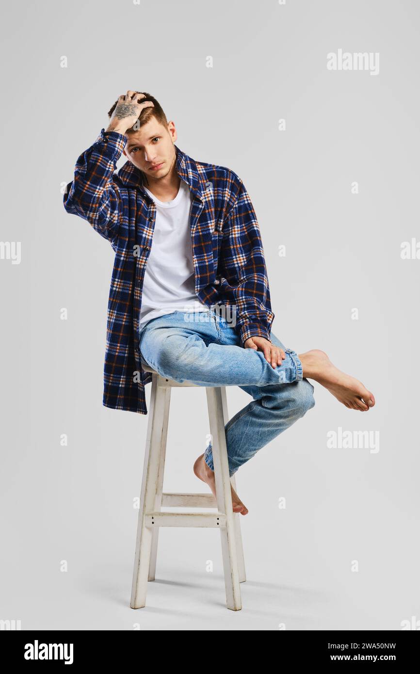 Full length studio portrait of young cocky man in shirt and jeans sitting on tall wooden chair Stock Photo