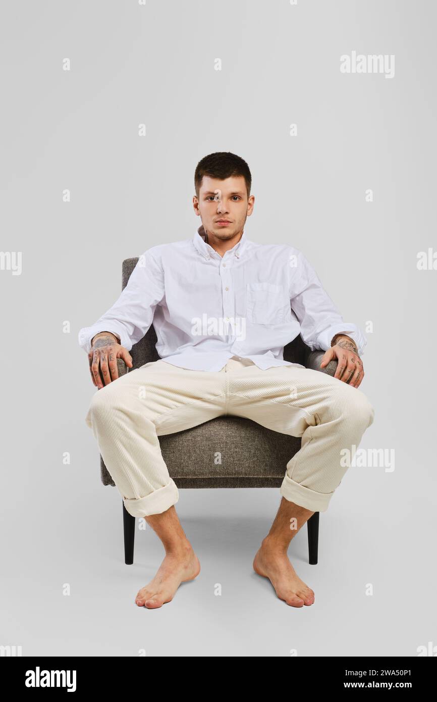 Full length studio portrait of young barefoot man in white shirt and ivory trousers sitting in armchair Stock Photo