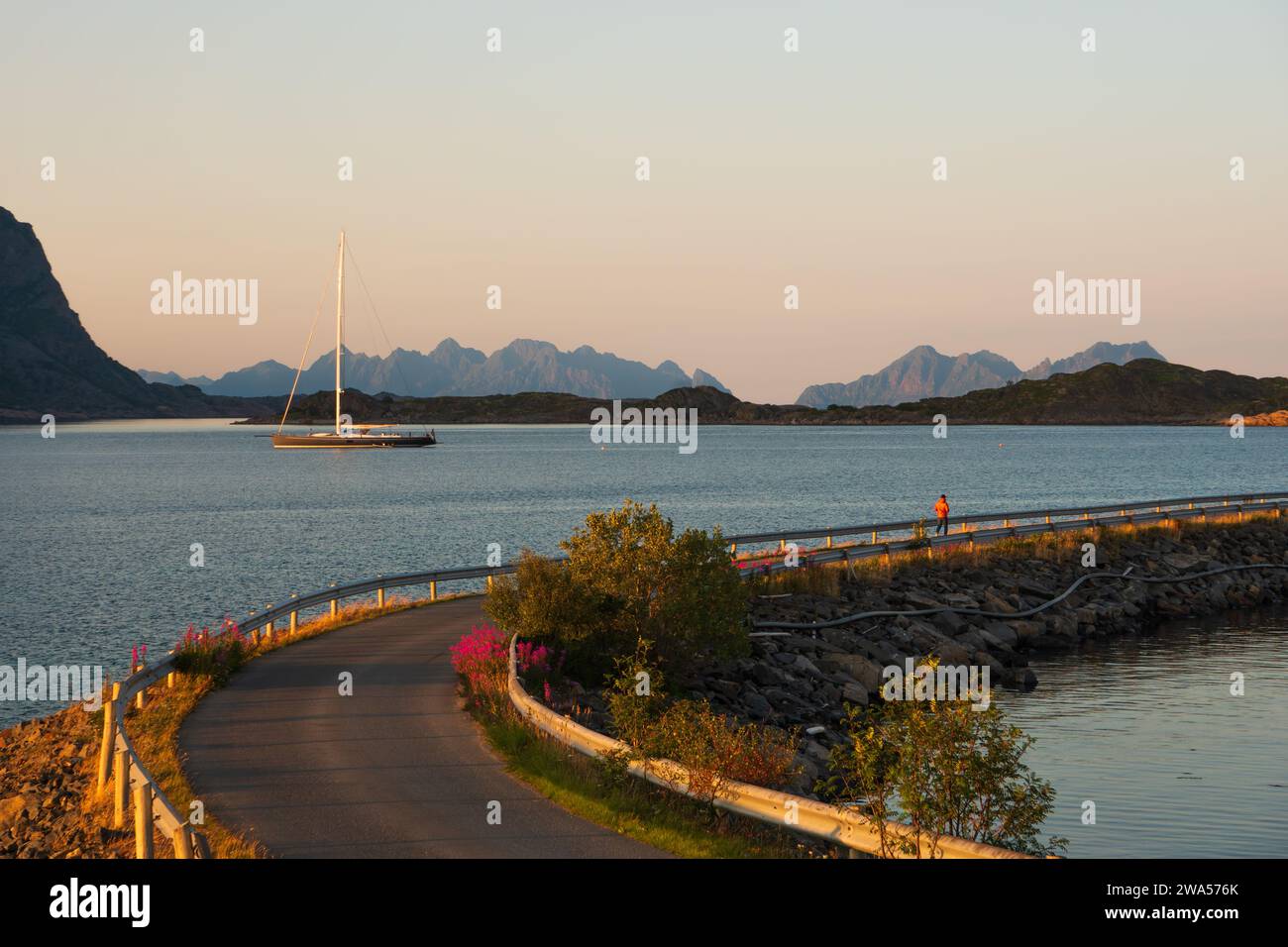 Sailboat near Henningsvaer, Lofoten, Norway in the midnight sun, man walking on a road in low sunlight. Sea and mountains, clear sunset sky. Stock Photo
