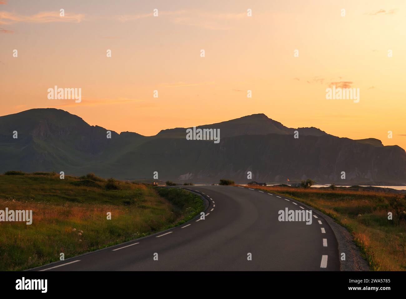 Midnight sunset over lofoten with a curved road heading towards the mountains, clear sky and deep orange blue colours. Green grass, soft colours. Stock Photo