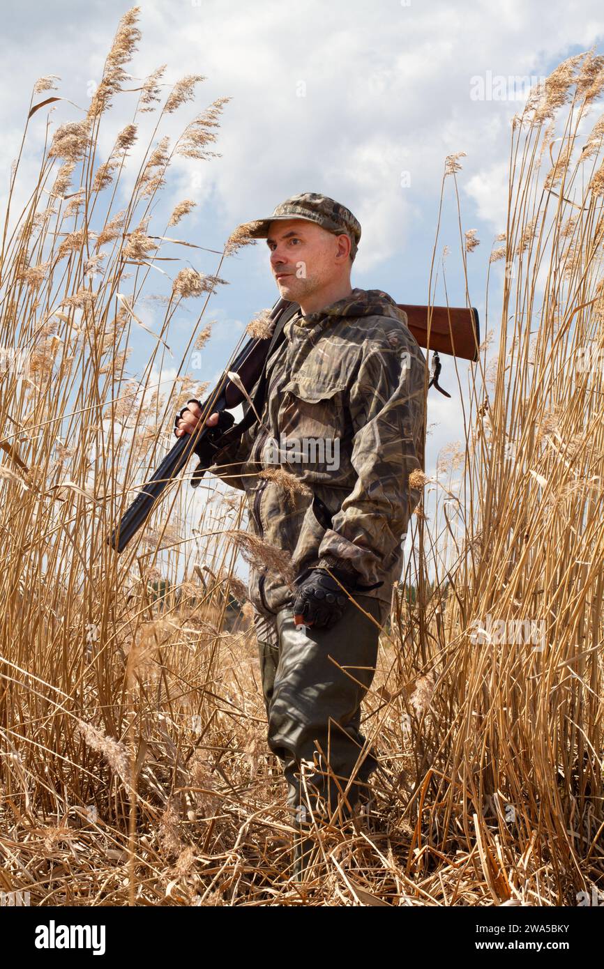 In late autumn, the grass on the shore of the lake turned yellow. The duck hunter holds a discharged shotgun on his shoulder. He stands among the tall Stock Photo