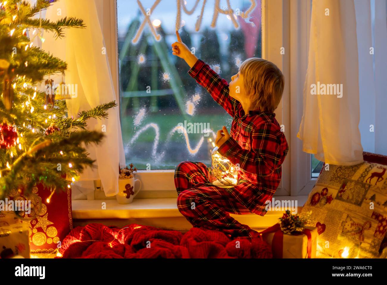 Cute child, sitting on a window, looking outdoors for Santa Claus, eating cookies Stock Photo
