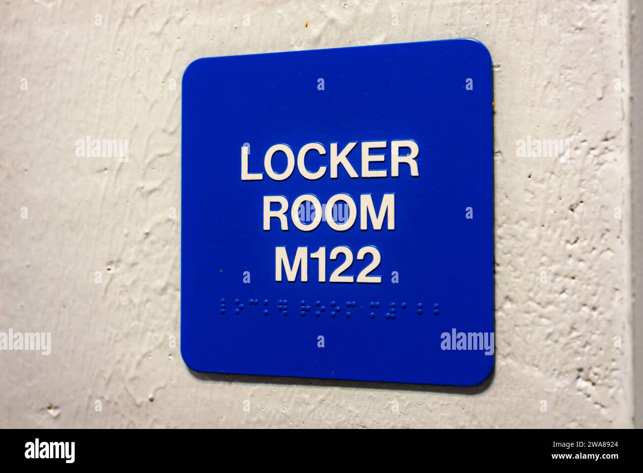 Blue LOCKER ROOM M122 sign with braille, on a white block wall. Stock Photo