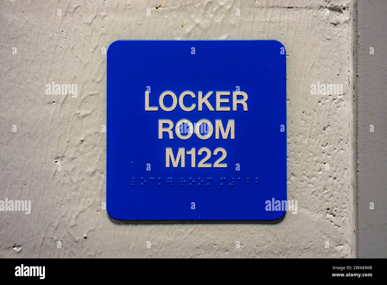 Blue LOCKER ROOM M122 sign with braille, on a white block wall. Stock Photo