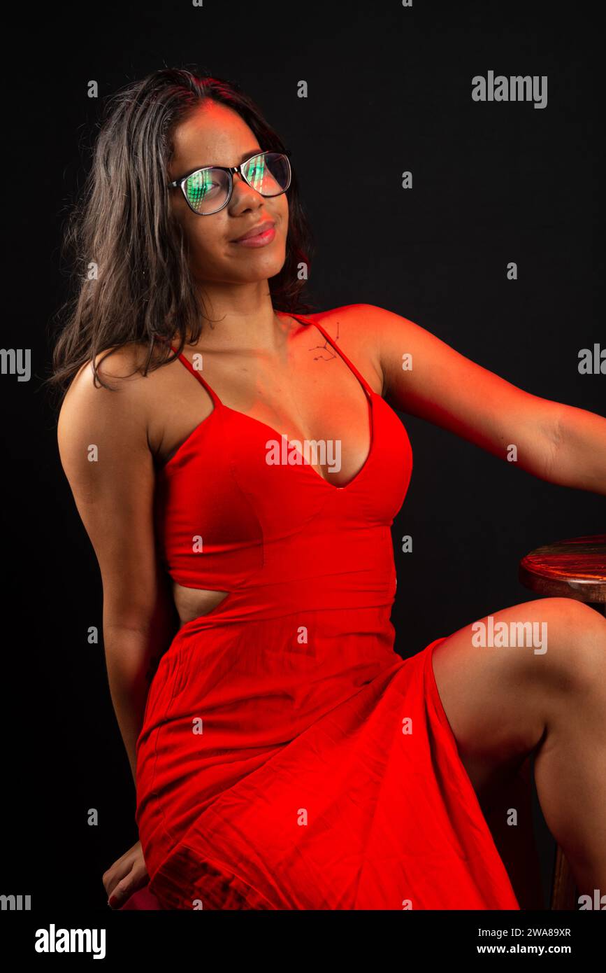 Portrait of beautiful young woman wearing glasses and in red dress sitting posing for photo. Isolated on black background. Stock Photo