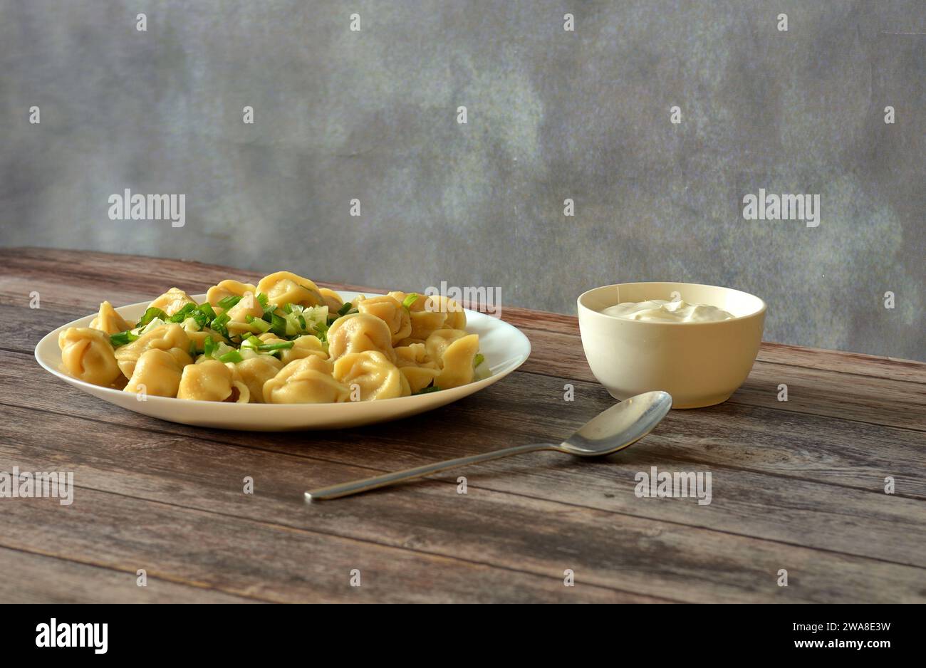 A plate with delicious freshly brewed dumplings sprinkled with chopped green onions on a wooden table, next to a spoon and a cup of sour cream. Close- Stock Photo