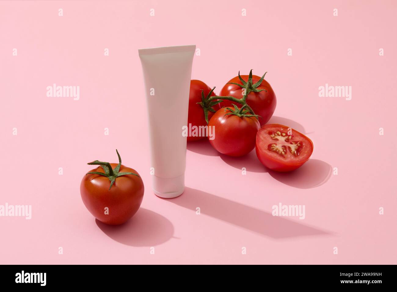 White tube without label decorated with several tomatoes. Pink background. Tomatoes are packed with enzymes that can exfoliate your skin gently Stock Photo