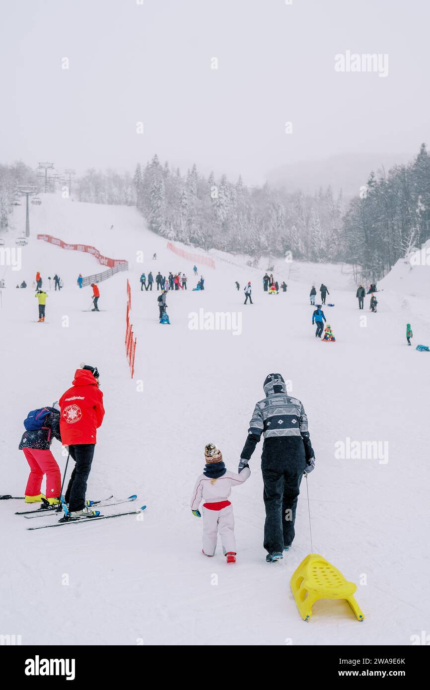 Mothers with children on skis and sleds climb a snow-covered ski slope ...