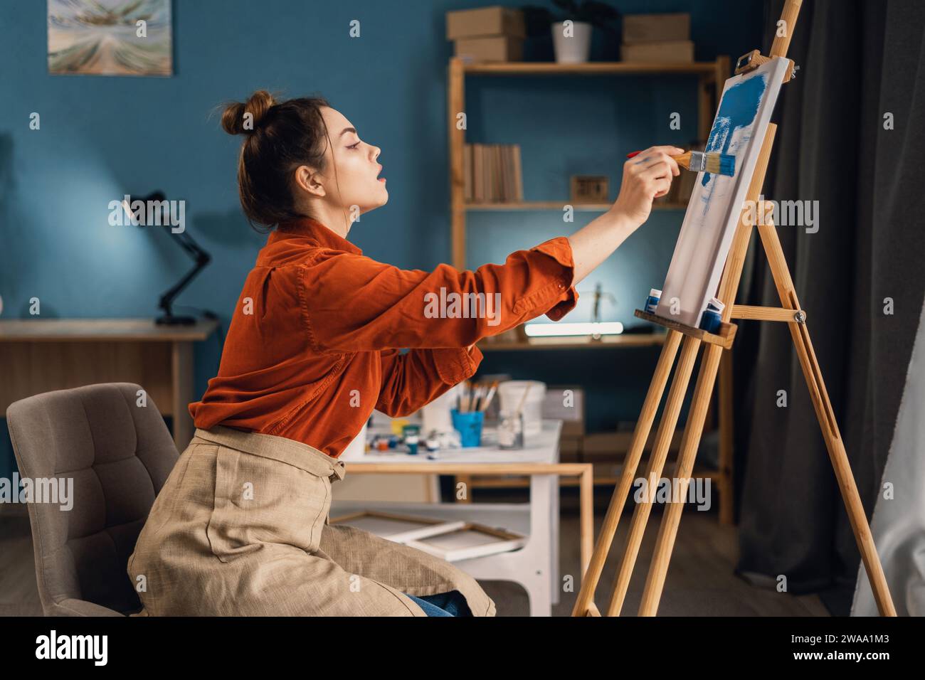 Young woman artist painting on canvas sitting at easel at home studio ...