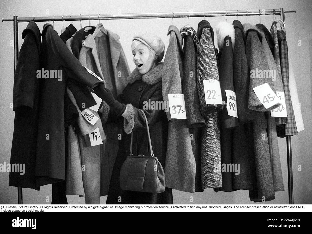 Great discounts of outerwear in the 1960s. A woman stands by a clothes rack, choosing from outerwear and coats marked with price tags with discounted prices. Judging by her face, she appreciates the reduced prices and thinks she's getting real bargains. Sweden January 1963. Ref CV28 Stock Photo