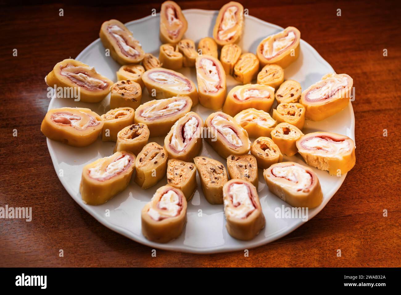 Sliced piece of cheese roulade with filling (ham, soft cheese, flax seed) on white plate on wooden table, closeup. Stock Photo