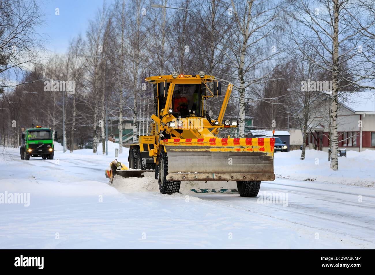 Yellow motor grader Veekmas FG 2327 S removing snow from street with adjustable blade, gritting lorry follows. Salo, Finland. December 27, 2023. Stock Photo