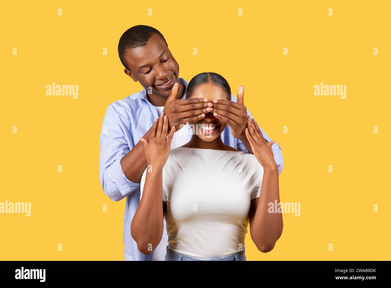 Black man covering woman's eyes with hands, both smiling on yellow Stock Photo