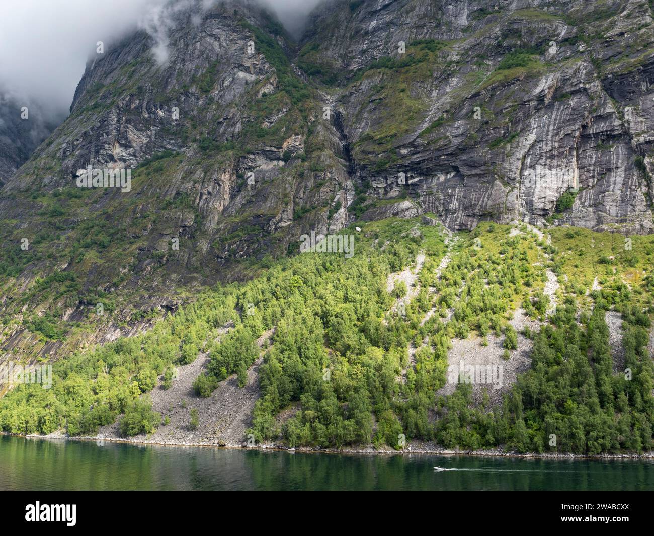 A tiny boat passing under a typical massive cliffs on the side of the Geirangerfjord near Geiranger, Norway. Stock Photo