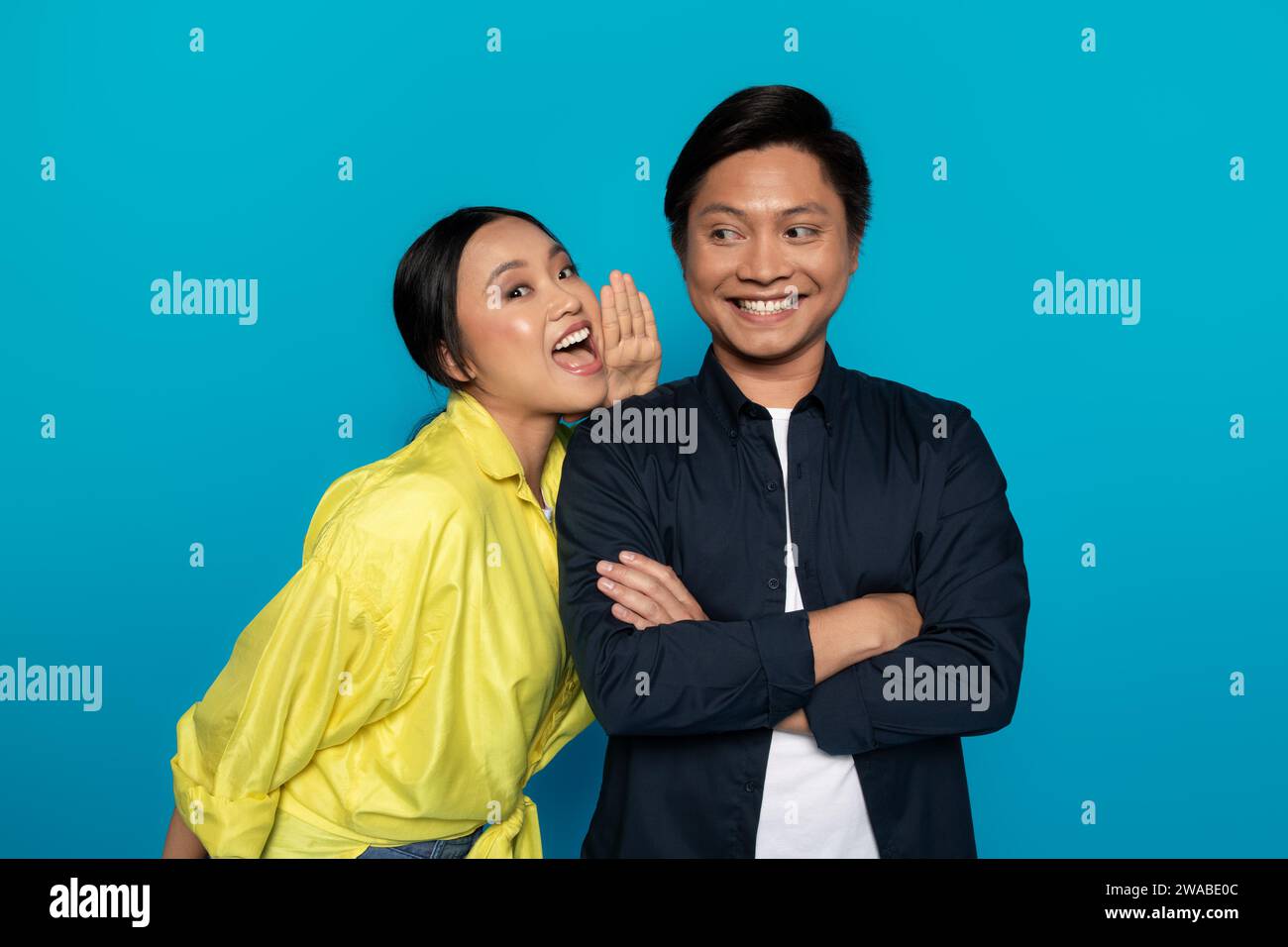 An Asian woman whispers into the ear of a smiling man with arms crossed, sharing a secret Stock Photo