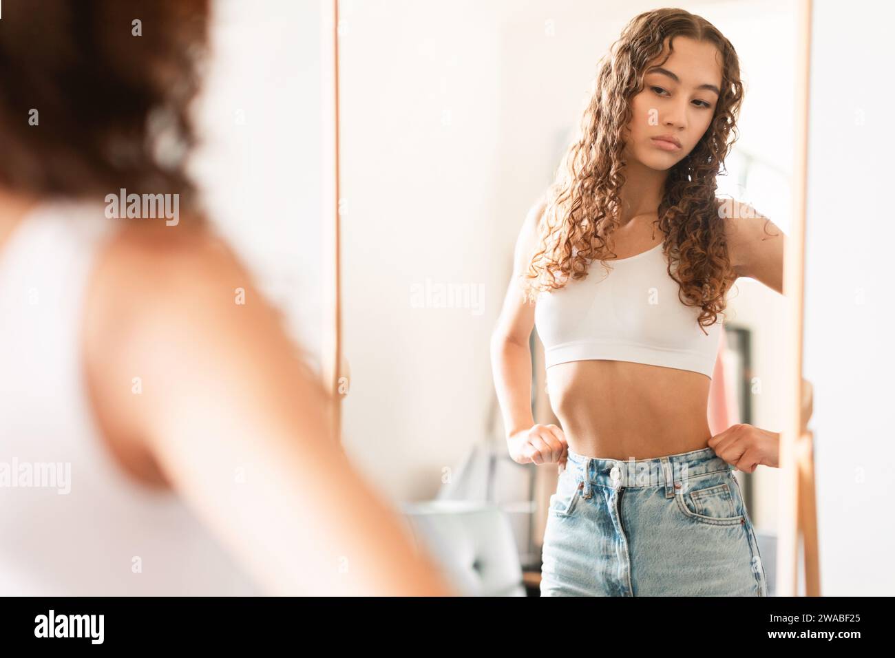 Unhappy teen girl looking critically at herself in mirror indoors Stock Photo