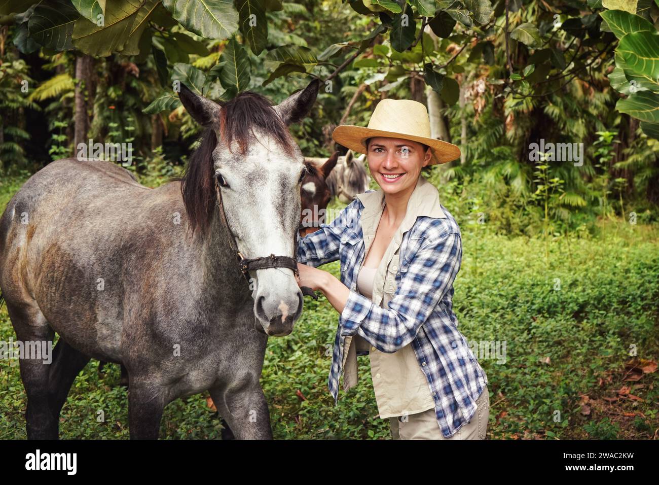 Young woman in shirt and straw hat posing next to gray horse, jungle trees background - horseriding, ranch at Isalo Park, Madagascar Stock Photo