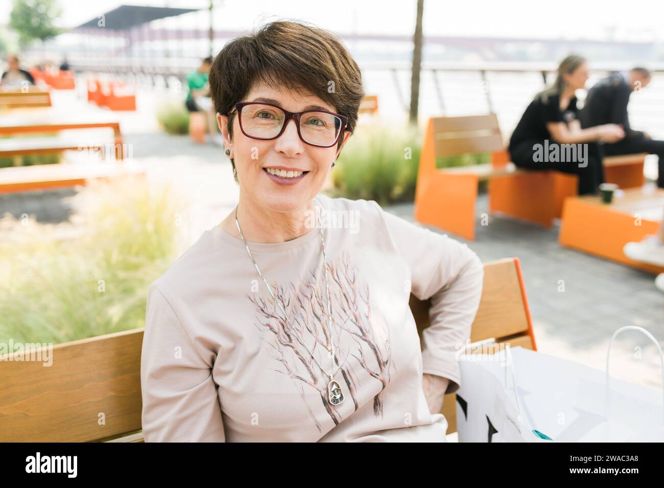 Outdoor portrait of beautiful and elegant middle age 55 - 60 year old woman wearing long sleeve top sitting on bench on the street. Copy space and Stock Photo